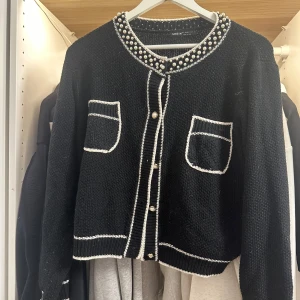 Svart kofta med pärlor från SHEIN - Snygg svart kofta från SHEIN med vita detaljer och pärlor längs kragen. Koftan har två fickor framtill, dekorativa knappar och är stickad i ett mjukt material. Perfekt för dig som gillar en chic och trendig look.