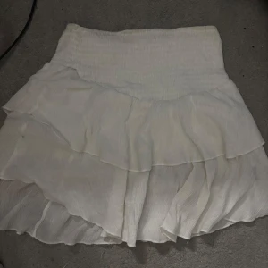 Vit volangkjol med resårmidja - Supersöt vit kortkjol med flera lager volanger och bred resår i midjan. Kjolen har en luftig och flowy känsla, perfekt för varma dagar. Passar dig som gillar en romantisk och feminin stil.