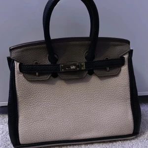 Elegant handväska i beige och svart - Snygg och stilren handväska i beige och svart med ett elegant lås framtill. 