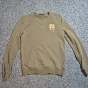 Beige sweatshirt från Morris Stockholm - Snygg beige sweatshirt från Morris Stockholm med broderad logga och brittisk flagga på bröstet. Tröjan har rund halsringning, långa ärmar och ribbade muddar. Perfekt för en chill och stilren look.