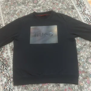 Svart sweatshirt från Hugo med tryck - Svart sweatshirt från Hugo med ett stort, skimrande logotryck på bröstet. Klassisk rund hals och långa ärmar. Tröjan är i mjuk bomull och har en clean, stilren look som passar perfekt till jeans eller joggers. Det finns ett litet hål på tröjan därför är priset så lågt.