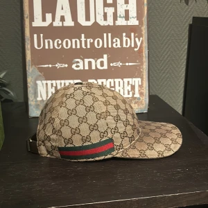 Gucci keps med GG-mönster - Snygg keps från Gucci i beige med klassiskt GG-monogram över hela kepsen. Kepsen har en böjd skärm och justerbart spänne bak. Detalj med grön och röd rand på sidan. Tillverkad i canvasmaterial med insida i svart tyg.