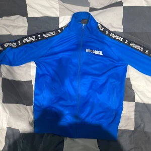Blå track jacket från Hoodrich M - Blå track jacket från Hoodrich med vit logga på bröstet och svarta band med Hoodrich-tryck längs ärmarna. Jackan har dragkedja framtill, hög krage och två fickor. Perfekt för en sportig och streetinspirerad look.