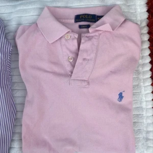 Rosa Ralph Lauren piké - Snygg och populär rosa pikétröja från Polo Ralph Lauren i slim fit-modell. Klassisk krage, knäppning med två knappar och blå broderad logga på bröstet. Perfekt för en clean och sportig look. Tillverkad i bomull för skön känsla. Nypris 1200kr.