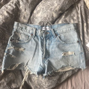  jeansshorts från Zara - Säljer ett par ljusblå jeansshorts från Zara med råa, fransiga kanter och slitningar framtill. Klassisk femficksmodell med hög midja och avslappnad passform. Perfekta för sommardagar när du vill ha en cool och avslappnad look.