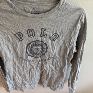 Grå långärmad tröja Polo Ralph Lauren - Snygg grå långärmad tröja från Polo Ralph Lauren med stort tryck framtill och klassisk rund hals. Tillverkad i mjuk bomull, perfekt för en avslappnad och stilren look. Logga även på ärmen för extra detalj.