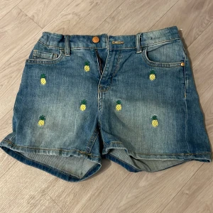 Jeansshorts med ananas från Lindex - Blå jeansshorts från Lindex med broderade små ananas över hela plagget. Klassisk femficksmodell med knapp och dragkedja framtill. Shortsen har normal passform och är tillverkade i bomull, perfekta för en chill sommardag.