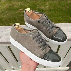 Grå sneakers från Lanvin med lackad tå - Snygga grå sneakers från Lanvin i mocka med cool lackad mörkgrå tå och vita sulor. Skorna har grå snörning och en stilren, minimalistisk design som passar perfekt till streetwear eller en clean look. Sulan är väldigt sliten men den kan man ersätta med en ny.