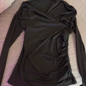 Svart omlott offshoulder topp - Snygg svart omlottopp med offshoulder-design och långa ärmar. Toppen har en figurnära passform och draperade detaljer längs sidorna som ger en cool och trendig look. Perfekt att styla med jeans eller kjol för en stilren outfit.