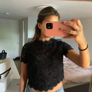 Svart spetsblus med dragkedja - Svart spets blus från ZARA med korta ärmar och rund hals. Blusen har ett detaljerat spetsmönster och en synlig dragkedja i ryggen. Croppad modell med vågig kant nertill 