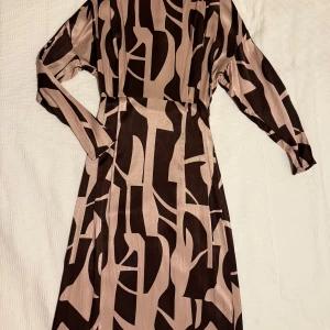 Mönstrad långklänning Stockh LM Studio - Snygg långklänning från Stockh LM Studio i glansigt material med grafiskt mönster i beige och mörkbrunt. Klänningen har lång ärm, markerad midja och dragkedja i sidan. Perfekt för dig som gillar statement-plagg med cool design. Helt oanvänd.