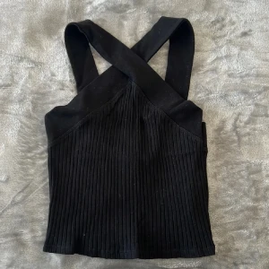 Svart ribbad korsettopp från Zara - Säljer en svart korsettopp från Zara med breda axelband som korsas i ryggen. Toppen är ribbad och har en croppad passform som ger en cool och modern look. Perfekt att styla med jeans eller kjol för en trendig outfit.