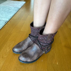 Bruna boots med stickad detalj - Snygga bruna boots med grov sula och stickad mudd upptill i vinröd ton. Skorna har dekorativa flätade remmar runt skaftet och en liten metallberlock. Perfekta för höst och vinter med sin robusta look och coola detaljer.