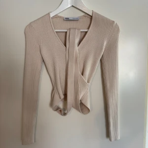 Offwhite ribbad omlottröja från Zara - Supersnygg offwhite ribbad omlottröja från Zara med långa ärmar och knytband i midjan. Tröjan har en v-ringad design och är gjord i mjukt stickat material som sitter skönt och ger en clean look. Lite kortare modell.