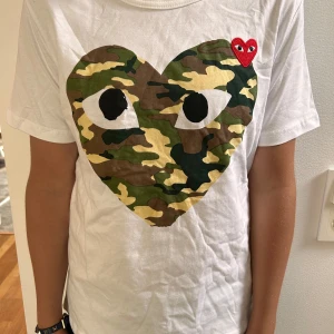 Comme des Garçons Play camo t-shirt - Vit t-shirt från Comme des Garçons Play med stort hjärta i camouflage-mönster och ögon på bröstet. Liten röd hjärtpatch med ögon uppe till höger. Klassisk rund hals och korta ärmar. Skön bomullskvalitet och snygg streetstil.