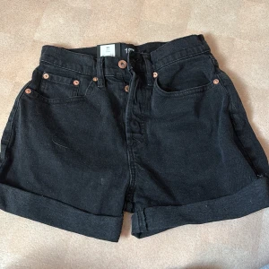 Svarta jeansshorts från 157 XS - Svarta jeansshorts från 157 i modellen DIBA. Högmidjade med klassisk femficksdesign, uppvikta benslut och snygga kopparfärgade nitar. Perfekta till sommaren och lätta att matcha med allt. Stilsäkra och bekväma shorts för dig som gillar en clean look.