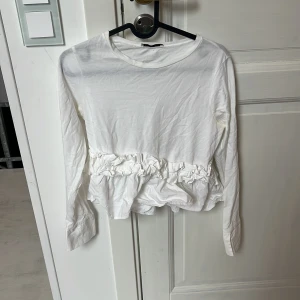 Vit långärmad blus med volang Zara - Supersöt vit långärmad blus från Zara med volangdetalj runt midjan. Den har en enkel rund halsringning och är gjord i mjuk bomull, perfekt för dig som gillar en clean och feminin stil.