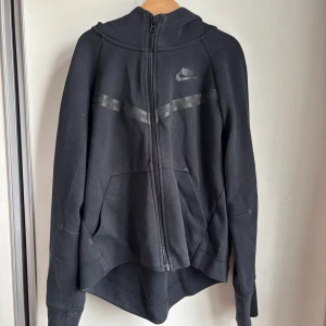 Svart Nike Tech hoodie med dragkedja - Svart hoodie från Nike med hel dragkedja och huva. Snygga svarta detaljer över bröstet och klassisk Nike-logga på vänster sida. Praktiska fickor framtill och mjukt bomullsmaterial. Perfekt för chill eller träning.