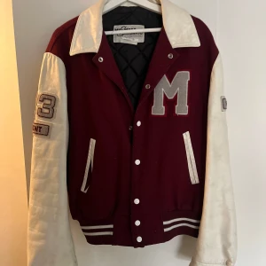 Vinröd och vit bomberjacka Mid-West - Snygg collegeinspirerad bomberjacka från Mid-West med vinröd kropp och vita ärmar. Stora broderade detaljer som ett grått M på bröstet och siffror på ärmen. Jackan har ribbade muddar, knappar framtill och två fickor. Perfekt för dig som gillar retrostil.