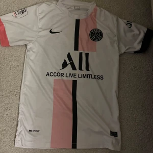 PSG Nike Dri-FIT fotbollströja 176 - Paris Saint-Germain matchtröja från Nike med Dri-FIT och VaporKnit-teknologi. Vit bas med rosa och svart vertikal rand, PSG-logga på bröstet och sponsortryck. Kortärmad och tillverkad i lätt, ventilerande material för maximal komfort på planen.