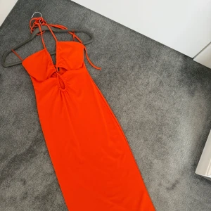 Orange långklänning med cut out perfekt för sommaren - Snygg orange långklänning med halterneck och smala axelband. Klänningen har en cool cut out-detalj framtill och är figurnära i modellen. Perfekt för dig som vill sticka ut med en färgstark look.