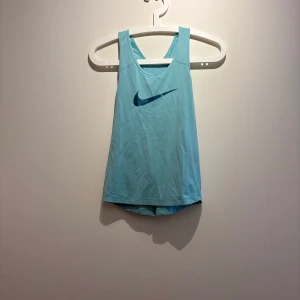Nike Dri-Fit blå linnetopp M - Sportig ljusblå linnetopp från Nike med Dri-Fit-teknologi. Toppen har en klassisk racerback, stort blått Nike-logga framtill och är tillverkad i ett lätt, ventilerande syntetmaterial som passar perfekt för träning.
