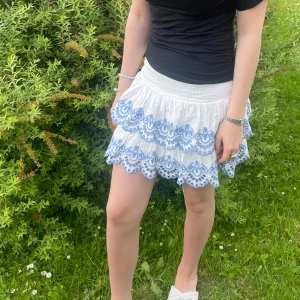 Vit volangkjol med blå broderi - Superfin vit kortkjol med flera volanger och blått broderi längs kanterna. Kjolen har smock i midjan för en bekväm passform och är perfekt för varma dagar. Den har en söt och somrig vibe med detaljerade broderier som verkligen sticker ut. Från lindex barn men passar xs/s