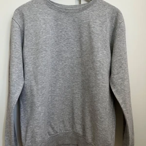 Grå sweatshirt från Kappahl - Enkel och stilren grå sweatshirt från Kappahl i storlek 158/164. Tröjan har rund halsringning, ribbade muddar vid ärmslut och nederkant samt lång ärm. Perfekt för en chill och avslappnad look.