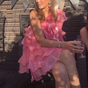 Rosa volangklänning oneshoulder - Superfin bubbelgumsrosa kortklänning med flera lager volanger och oneshoulder-design. Klänningen har en glansig finish och är perfekt för dig som vill sticka ut. Snyggt flowy material och riktigt statement-plagg för sommarens alla tillfällen och temafester! (Nypris 1250kr)