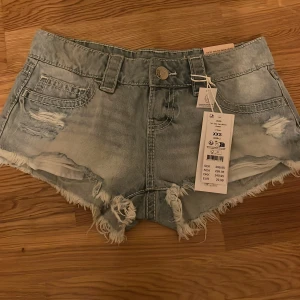 Lågmidjade jeansshorts Gina Tricot - Snygga ljusblå lågmidjade jeansshorts från Gina Tricot med slitna detaljer, fransig kant och coola nitar på bakfickorna. Perfekta för sommaren och ger en avslappnad vibe. Klassisk femficksmodell med knapp och dragkedja framtill. Nyskick med prislapp!💕