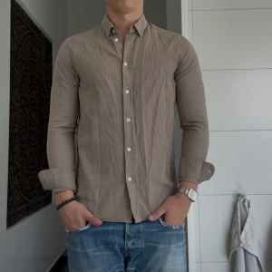 Beige skjorta från Samsøe Samsøe - Stilren beige skjorta från Samsøe Samsøe med klassisk krage och vita knappar framtill. Skjortan är långärmad och har en smal passform som sitter snyggt på kroppen. Perfekt till jeans för en clean look.