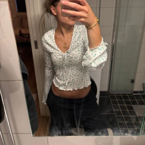 Vit blommig croppad blus med knyt - Supersöt vit blus med små blå blommor, croppad modell och v-ringning. Långa ärmar med volangkant och ribbad struktur. Söt knytning framtill och vågig kant nertill ger en romantisk vibe. Perfekt för vår och sommar!