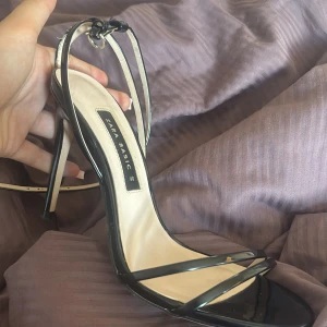 Svarta klackar från Zara  - Snygga svarta pumps från Zara Basic med smala remmar och hög klack. Skorna har öppen tå och justerbar rem runt vristen. Tillverkade i syntetmaterial med beige innersula. Perfekta för dig som vill ha en stilren och elegant look.