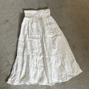Vit långkjol i bomull med smock - Superfin vit långkjol i luftig bomull med bred smock i midjan för en bekväm passform. Kjolen har volangdetaljer och ett flowy fall som ger en chill och somrig vibe. Perfekt att styla med crop top eller linne för en avslappnad look.