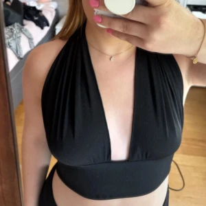 Svart ribbad knyt-topp med djup v-ringning - Supersnygg svart ribbad topp med bred midjeresår och djup v-ringning. Toppen knyts i nacken för en halterneck-look och är croppad. Perfekt för dig som vill sticka ut med en trendig och stilren vibe. Materialet är stretchigt och mjukt mot huden. Går att knyta på olika sätt. Storlek S