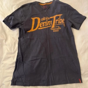 Mörkblå t-shirt EDC  - Snygg mörkblå t-shirt från EDC med stor gul print 'Denim Tribe' på bröstet. Klassisk v-ringad modell med korta ärmar och normal passform. Perfekt för dig som gillar streetwear och coola tryck.