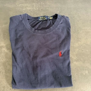 Mörkblå t-shirt från Polo Ralph Lauren - Snygg mörkblå t-shirt från Polo Ralph Lauren med klassisk röd broderad logga på bröstet. T-shirten har rund halsringning och är tillverkad i mjuk bomull. Perfekt för en clean och stilren look.