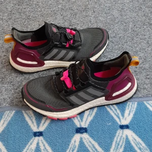 Adidas Ultraboost svarta sneakers - Säljer ett par Adidas Ultraboost sneakers i svart med mörklila och rosa detaljer. Skorna har gråa ränder på sidorna, vit platt sula och cool snörning med rosa snörlås. Ovandelen är i syntetmaterial och mesh för extra komfort. Perfekt för dig som gillar sportig stil.