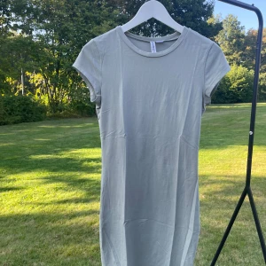 Grågrön t-shirtklänning från H&M  - En enkel och stilren grågrön t-shirtklänning från H&M. Klänningen har rund halsringning, korta ärmar och rak passform. Perfekt för dig som gillar minimalistisk och avslappnad stil. Materialet är mjuk bomull som känns skönt mot huden.