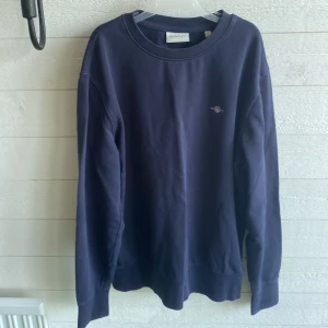 Mörkblå sweatshirt från GANT - Snygg mörkblå sweatshirt från GANT med klassisk rund hals och diskret logga broderad på bröstet. Tröjan har långa ärmar och är gjord i mjuk bomull, perfekt tröja för alla årstider. Passar dig som gillar enkel och stilren design.