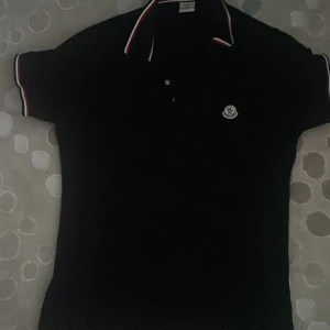 Svart Moncler pikétröja med röd/vit kant - Svart pikétröja från Moncler med klassisk krage och korta ärmar. Tröjan har vita och röda ränder på krage och ärmslut samt Moncler-logga på bröstet. Snygg och stilren modell i mjuk bomull, perfekt för dig som gillar exklusiva märken och sportig vibe.