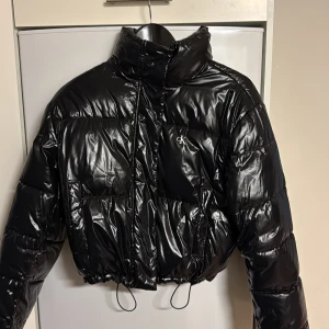 Svart pufferjacka från Calvin Klein - Snygg svart pufferjacka från Calvin Klein Jeans med glansig finish och croppad passform. Jackan har hög krage, dragkedja framtill, två stora fickor och justerbar snörning i nederkant. Perfekt för dig som vill ha en trendig och varm jacka med streetkänsla.
