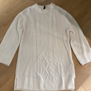 Vit stickad klänning  - Säljer en vit stickad klänning med snyggt kabelmönster framtill och grovstickade sidor. klänningen har lång ärm, ribbade muddar och en högre hals som ger en chill vibe. Perfekt för att hålla sig varm och se fräsch ut under hösten och vintern.