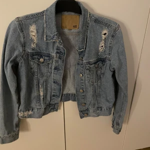 Ljusblå jeansjacka med slitningar 157 - Snygg ljusblå jeansjacka från 157 med slitna detaljer och klassiska bröstfickor. Jackan har knappar framtill och långa ärmar. Perfekt för dig som gillar en avslappnad och trendig look.