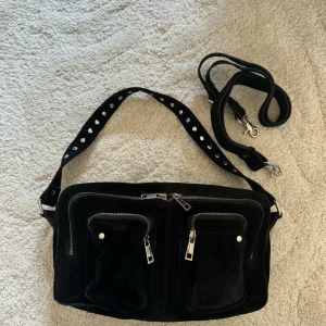 Svart mocka axelväska från Nunoo - Cool svart axelväska i mocka från Núnoo med flera fickor och silvriga dragkedjor. Väskan har både bred axelrem med nitar och en extra rem, perfekt för att bära crossbody. Snygg och praktisk med detaljer som ger en edgy vibe.Väska från Nunoo modell Ellie, större varianten. Knappt använd, mycket bra skick. Enda skavanken jag kan se är pyttelite missfärgat på 2 av dragkedjorna, var god se sista bilden. Kan användas på axeln eller som crossbody.