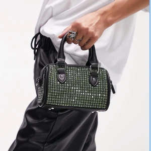 Svart handväska med gröna stenar - Obs första bilden är inte min! Topshop – Catlin - Svart bowler väska med grön strass. Cool svart handväska med glittriga gröna stenar på hela väskan. Väskan har både handtag och avtagbar axelrem, samt dragkedja upptill. Rektangulär form och detaljer i silverfärgad metall. Perfekt för dig som vill sticka ut. Jättefin bowler väska som tyvärr aldrig kommit till användning. Nypris 500. Jag har massor av artiklar till salu! 