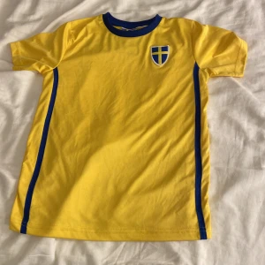 Svensk gul fotbollströja 134/140 - Gul fotbollströja med blå detaljer och svensk flagga på bröstet. Blå ränder längs sidorna och blå krage. SWEDEN tryckt på ryggen. Tillverkad i mjukt, lätt material som passar perfekt för träning eller match.