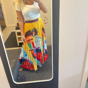 Färgglad plisserad långkjol med tryck - Säljer en maxilång plisserad kjol med färgstarkt tryck i gult, rött, blått och lila. Kjolen har bred lila midjeresår och coolt motiv med ansikten och grafiska former. Perfekt statement piece för dig som vill sticka ut.Köpt i Italien i juni 2025.Helt ny