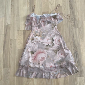 Blommig ljus kortklänning med volang - Superfin kortklänning med blommigt mönster i ljusa toner av rosa, beige och vitt. Klänningen har tunna axelband, volang upptill och längs nederkanten, samt en lätt A-linjeformad siluett. Perfekt för sommarens alla tillfällen!