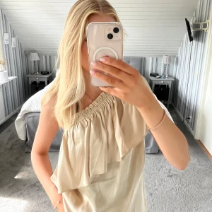 Guldig/Beige oneshoulder topp med volang - Säljer en guldig/beige oneshoulder topp med snygg volangdetalj framtill. Toppen har en avslappnad passform och ärmlös design med rynkad kant över axeln. Perfekt för dig som gillar en stilren och trendig look. Den är köpt vintage så vet faktiskt inte vart den är ifrån från början🩷
